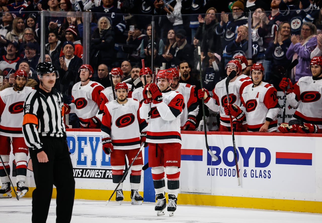 A hockey player in white and red brings his hands together as his teammates on the bench and the audience in the stands rise to applaud him.