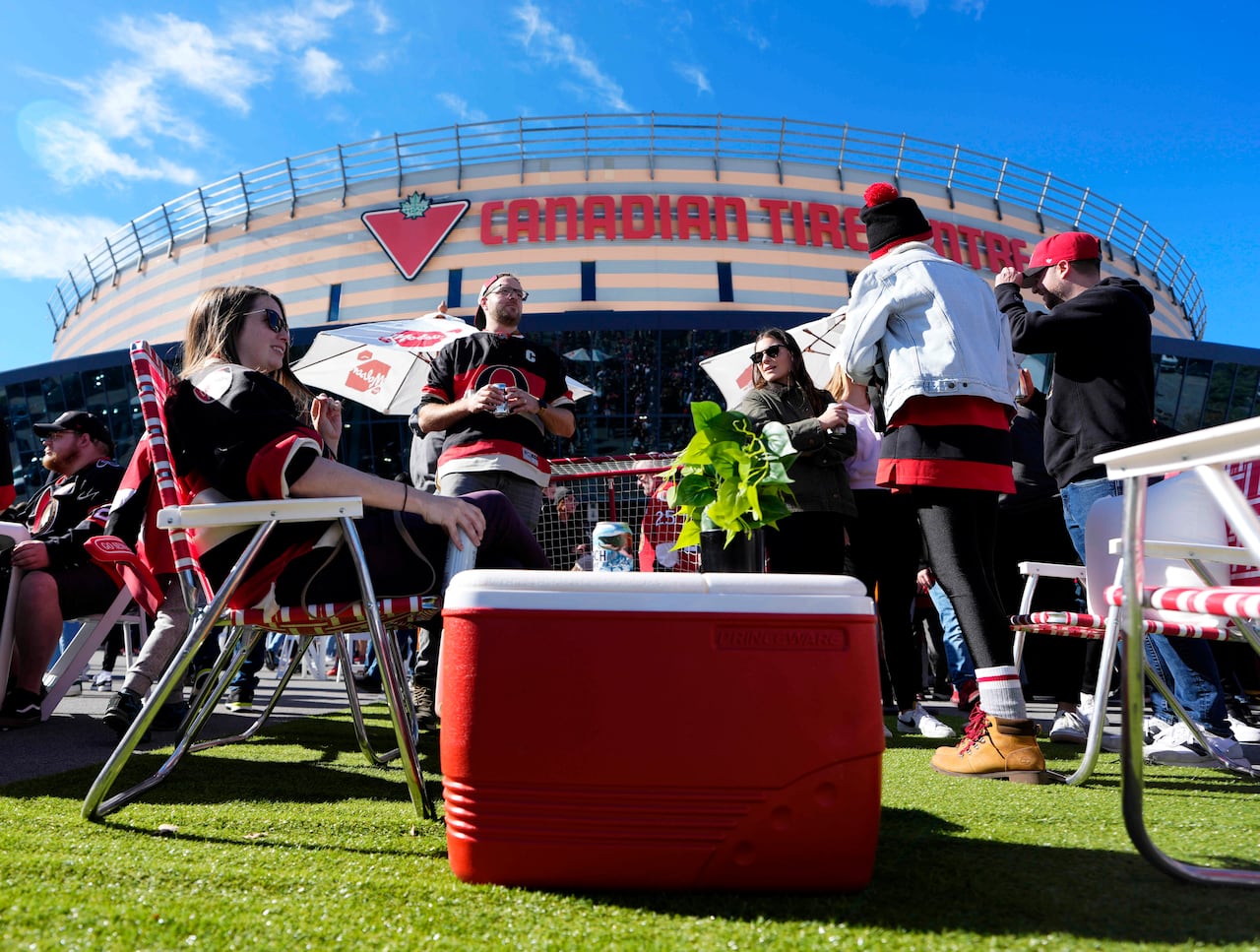 Several people wearing hockey jerseys gather outside a hockey arena on a warm day. A cooler is in the foreground.