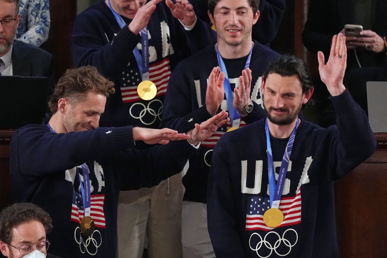 A man sporting a gold medal around his neck beams and lifts his arm while nearby individuals stand and applaud.