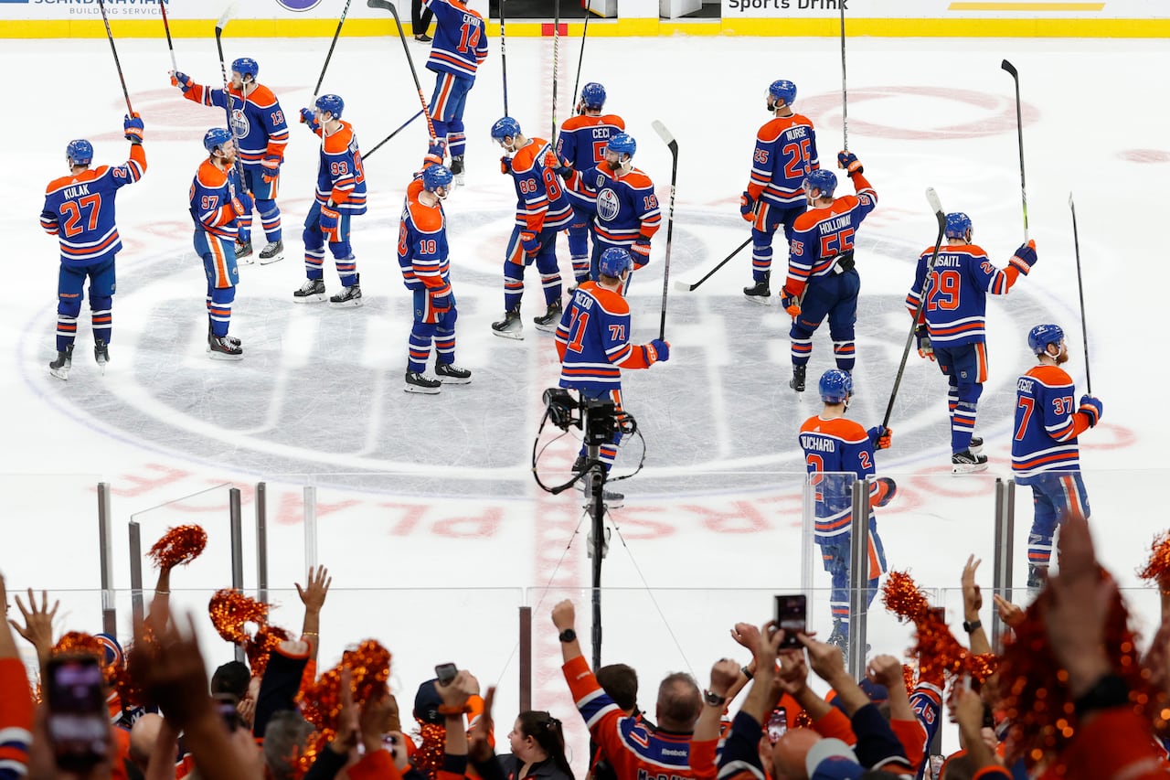 Hockey players on an ice rink raise their sticks in a gesture to their supporters as people in the crowd cheer.