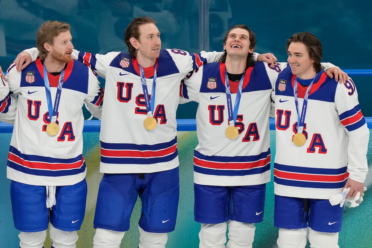 Four cheerful hockey players wearing USA jerseys stand linked arm-in-arm with gold medals around their necks.