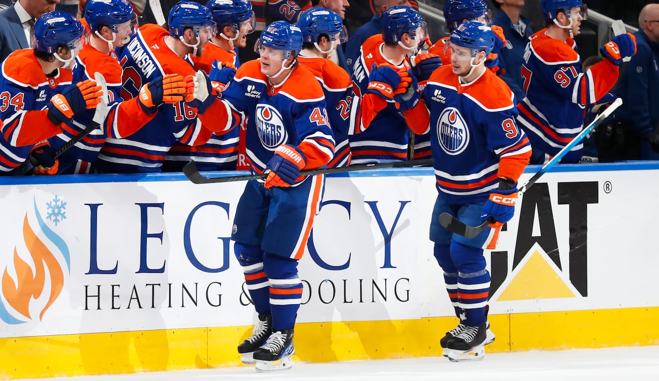 Two men's hockey players in blue-and-orange uniforms are fist-bumping their teammates on the bench after scoring a goal.