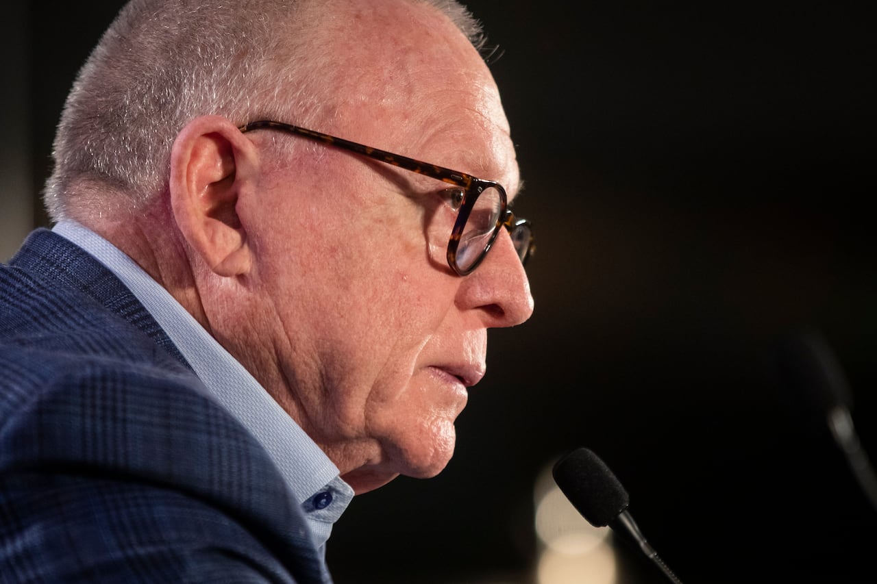 A man with glasses and a blue top is shown from the side, addressing a microphone.