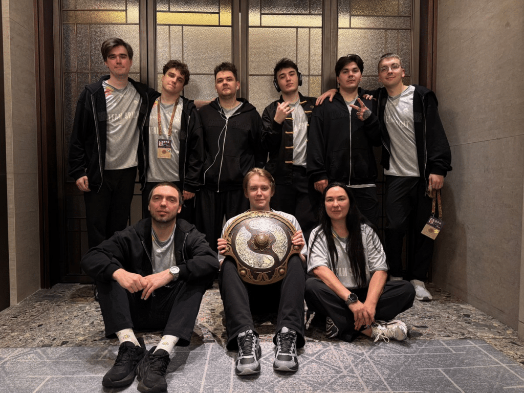 The image shows Team Spirit posing together in front of a modern wood-paneled wall, celebrating their victory at The International 2026 in China.