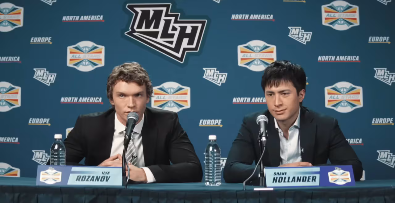 A pair of men participate in a media briefing. They are clad in dark suit jackets over white collared shirts, with a background featuring multiple promotional emblems.