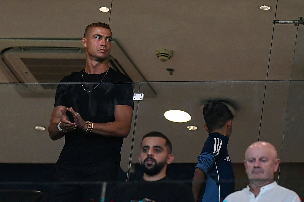 Cristiano Ronaldo stood with his son, looking away from the camera. He is in a box at a football game, two spectators are sat in front of him.