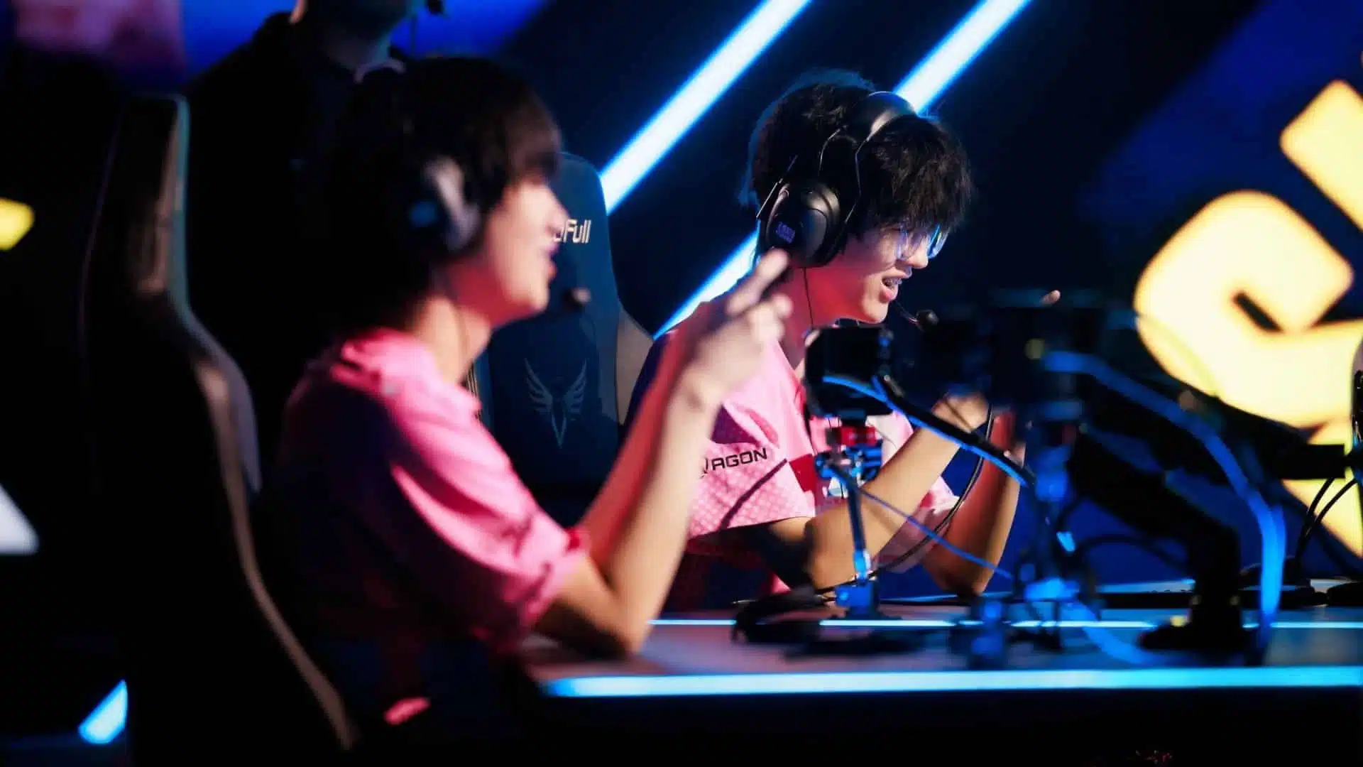 Two professional esports players in pink jerseys wearing headsets at a competition stage.